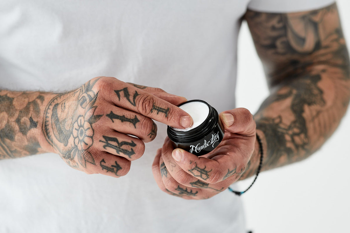 Tattooed hands holding an open black jar of white cream against a white shirt